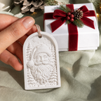 White paintable MelodyCast Santa Christmas ornament shown by hand with a holiday gift